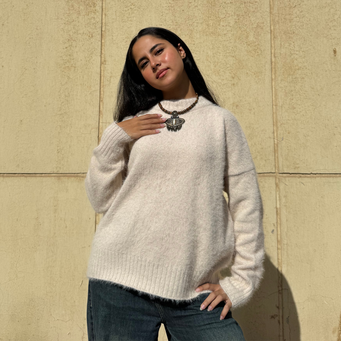 Woman wearing a beige sweater and blue jeans against a beige wall
