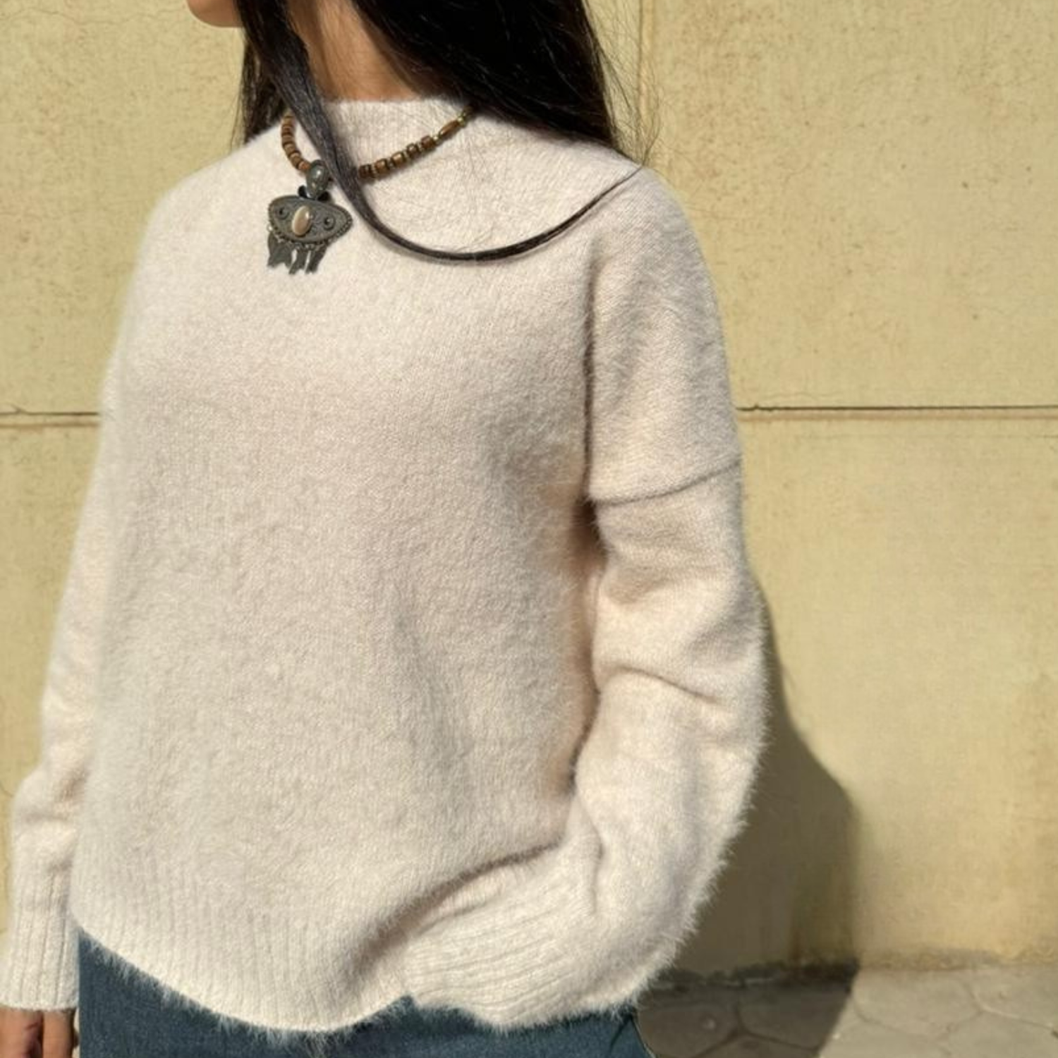 Woman wearing a beige sweater and blue jeans against a beige wall