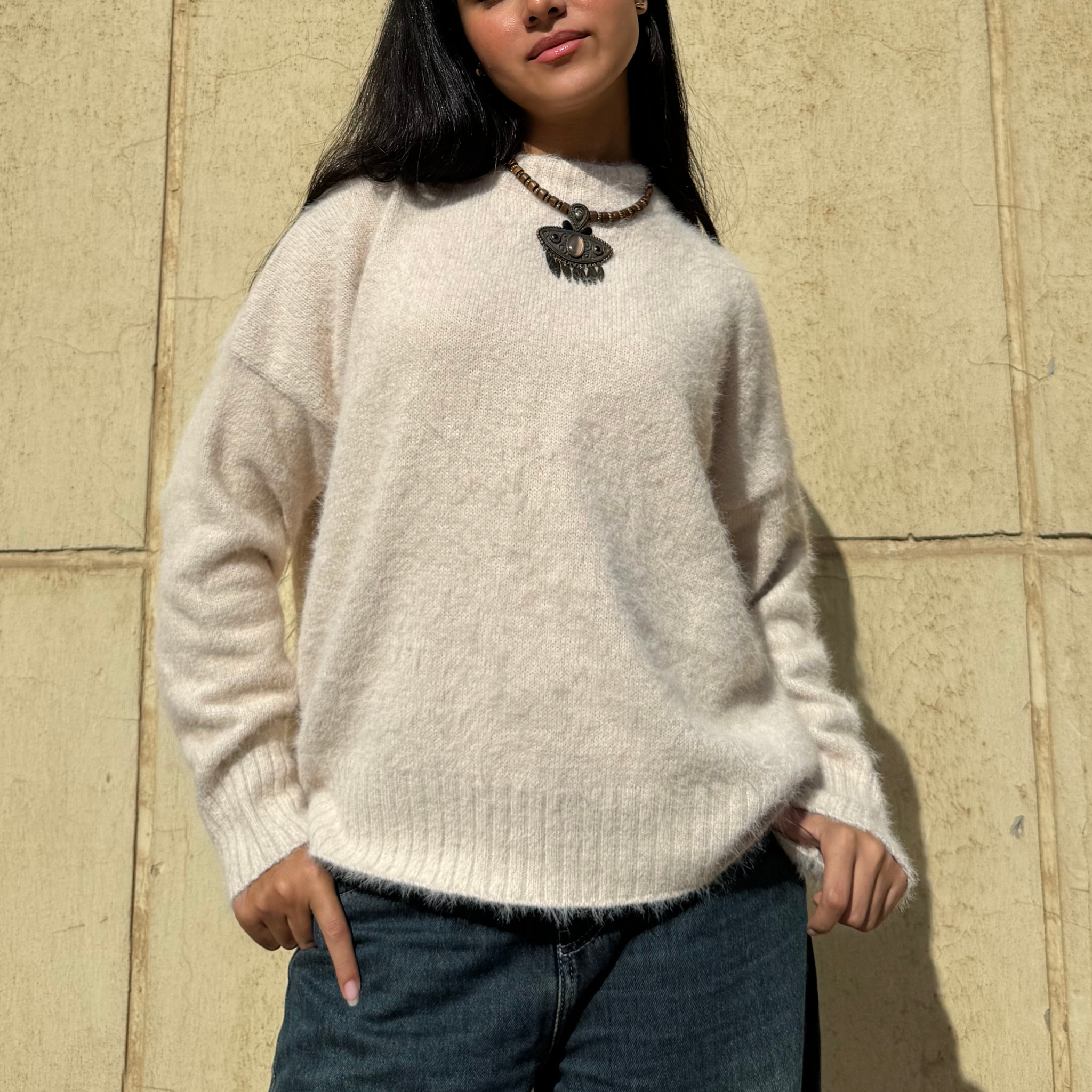 Person wearing a beige sweater and dark jeans against a beige wall