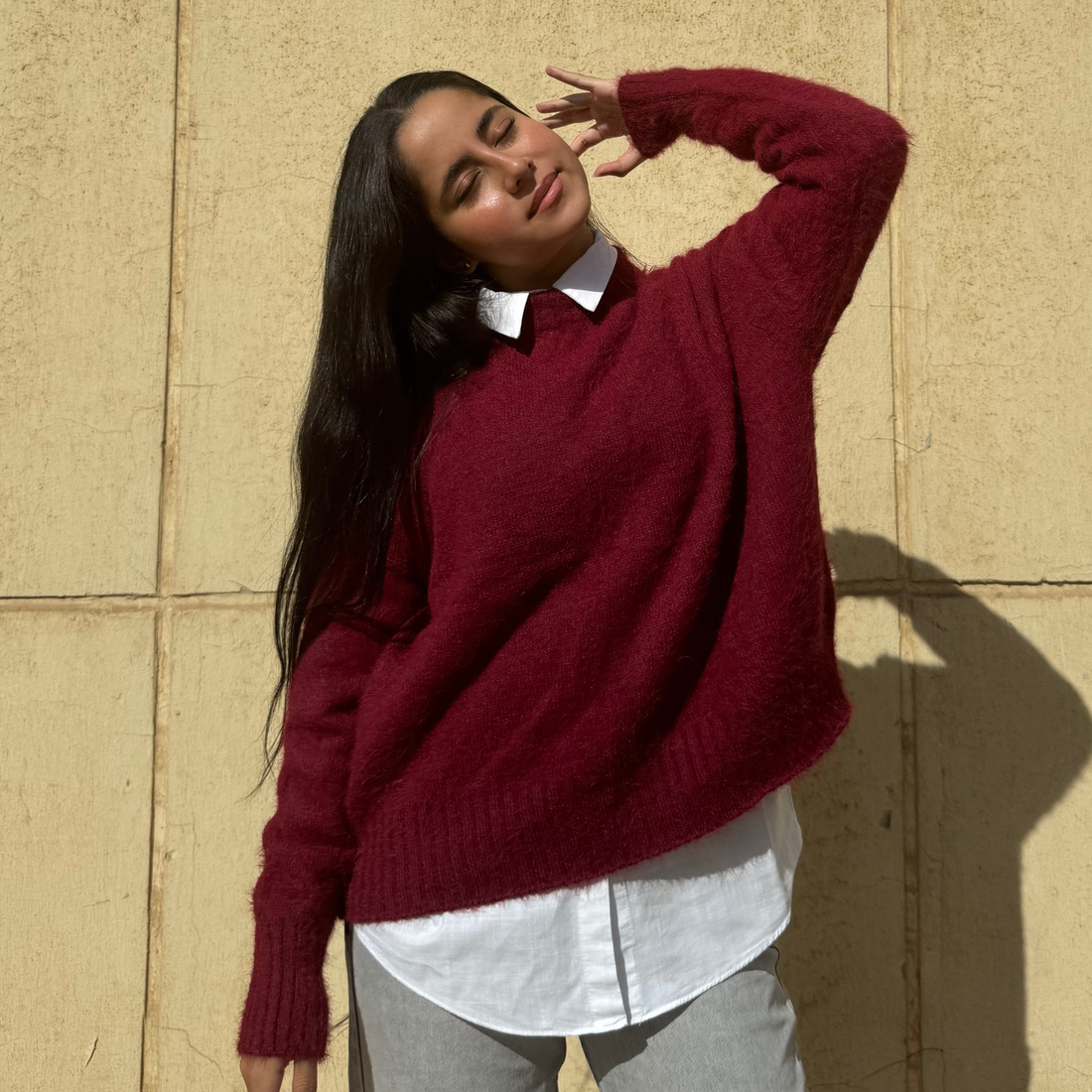 Woman wearing a Burgundy sweater over a white shirt against a beige wall.