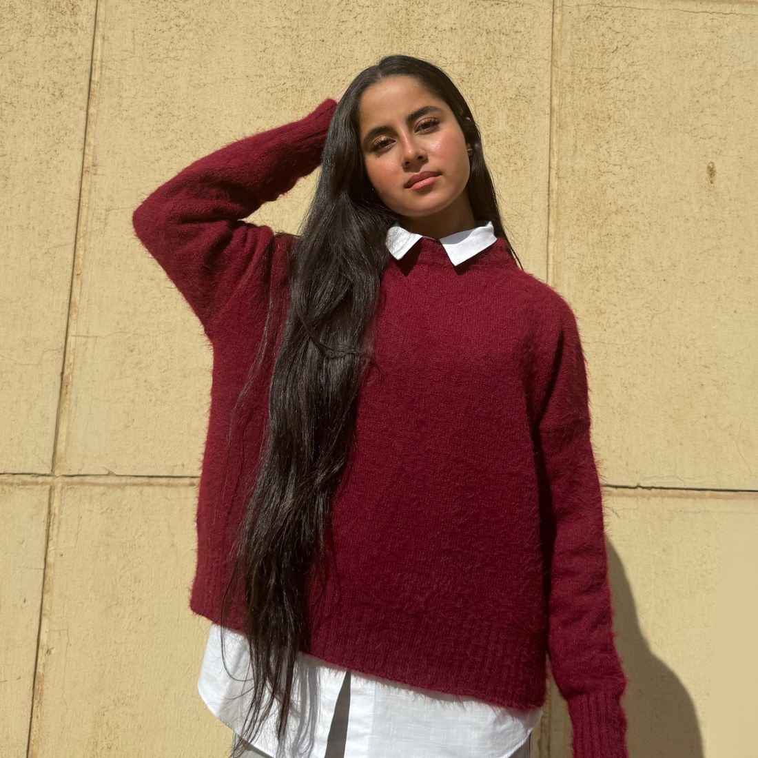 Woman wearing a Burgundy sweater over a white shirt against a beige wall.
